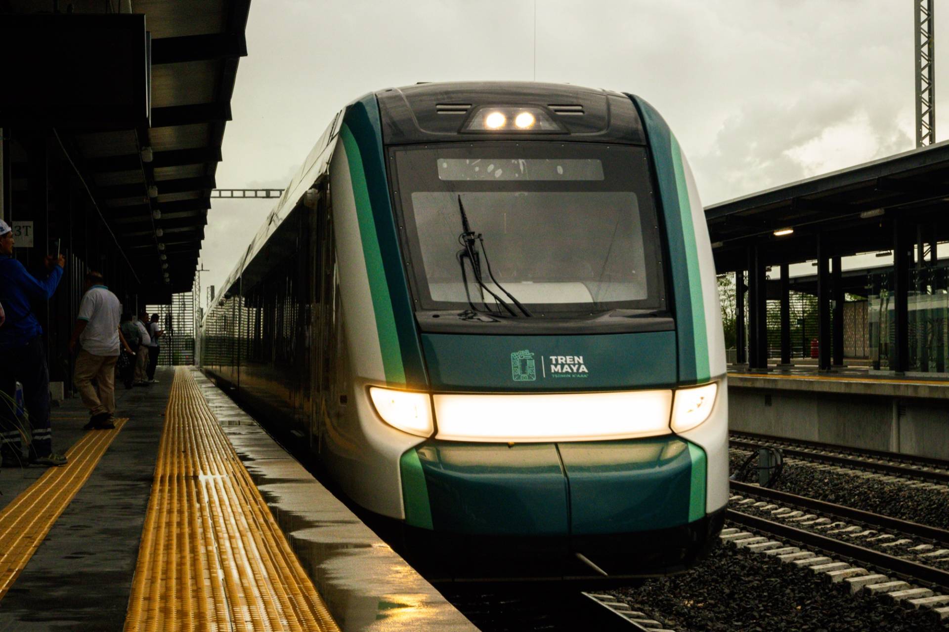 A green-and-white Tren Maya train with its headlights on arriving at the platform of Mérida Teya station under an overcast sky, with waiting passengers taking photos from the platform edge