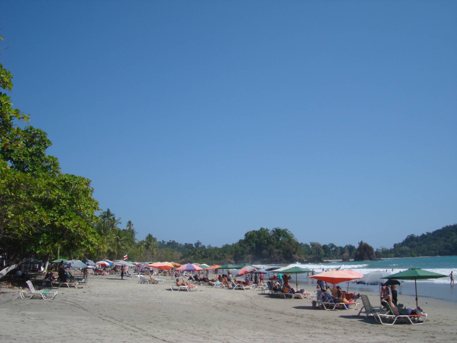 Rows of colourful sun umbrellas and white loungers on a wide sandy beach at Manuel Antonio, with almond trees on the left and a forested headland behind the surf