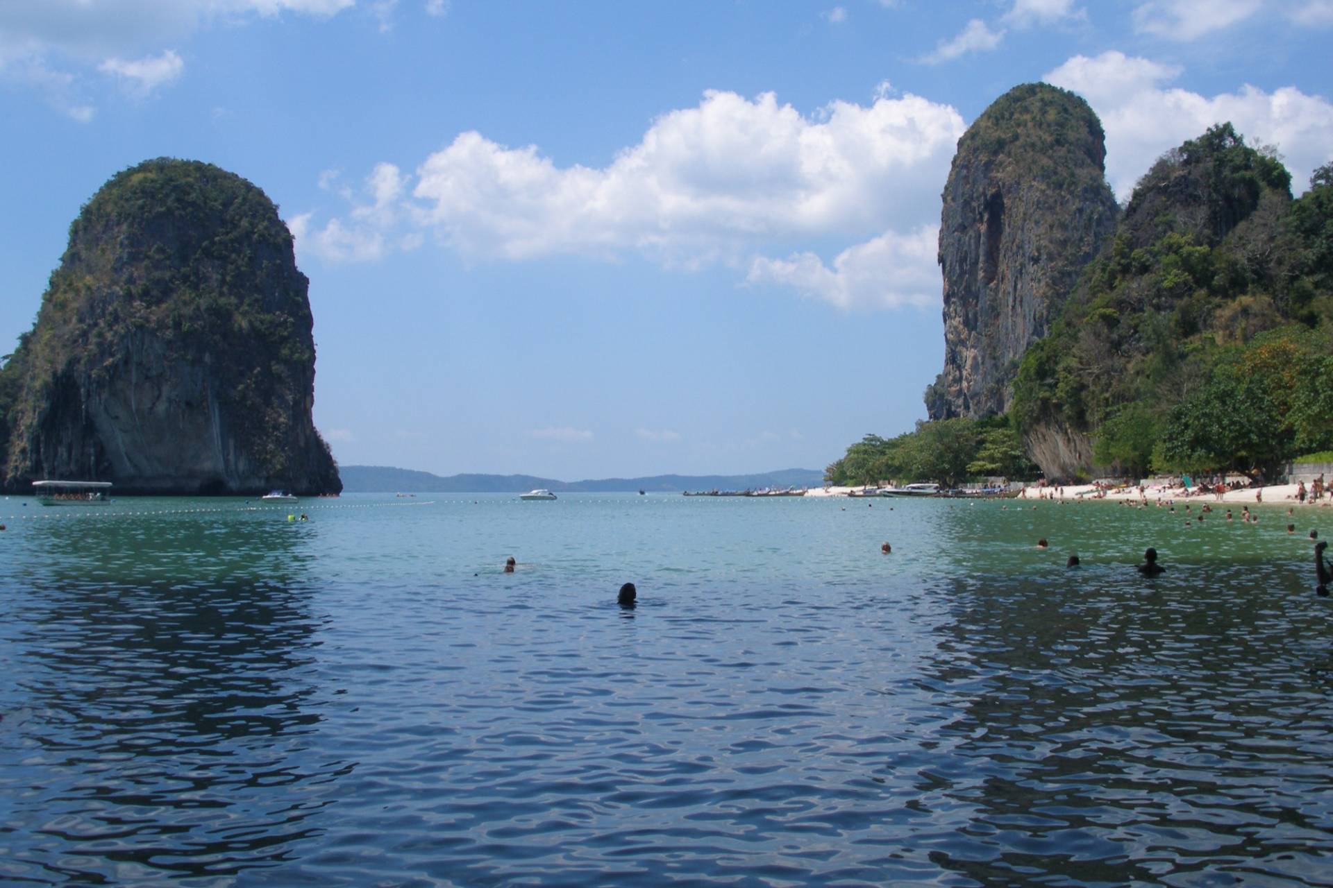 Railay Beach, Krabi: a calm bay with swimmers and moored boats between two tall limestone karst towers under a partly cloudy sky, with a strip of white sand and trees on the right