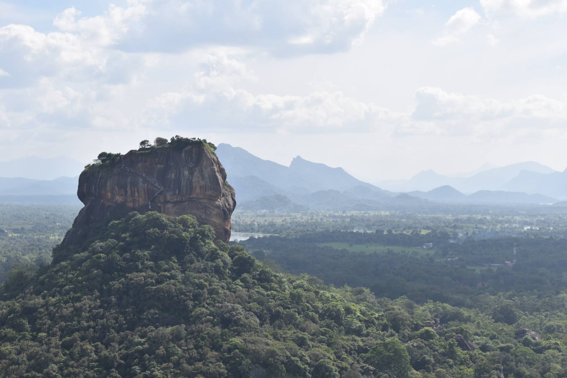 Sigiriya rock fortress seen from Pidurangala: a flat-topped orange rock with the staircase visible on its side, rising above forest with blue hills on the horizon