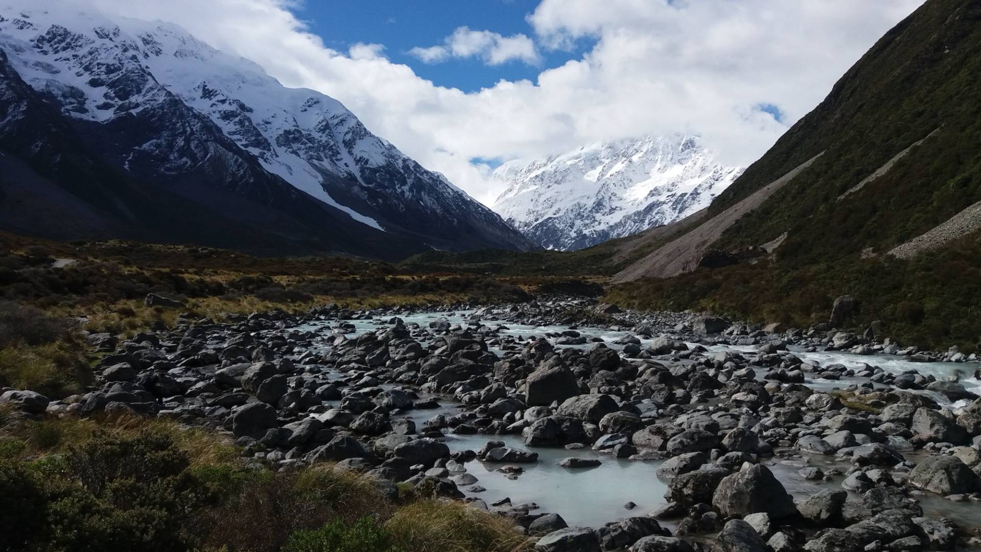 Rocky bed of the Hooker River on the Hooker Valley Track with snow-covered peaks of Aoraki/Mount Cook National Park under broken cloud