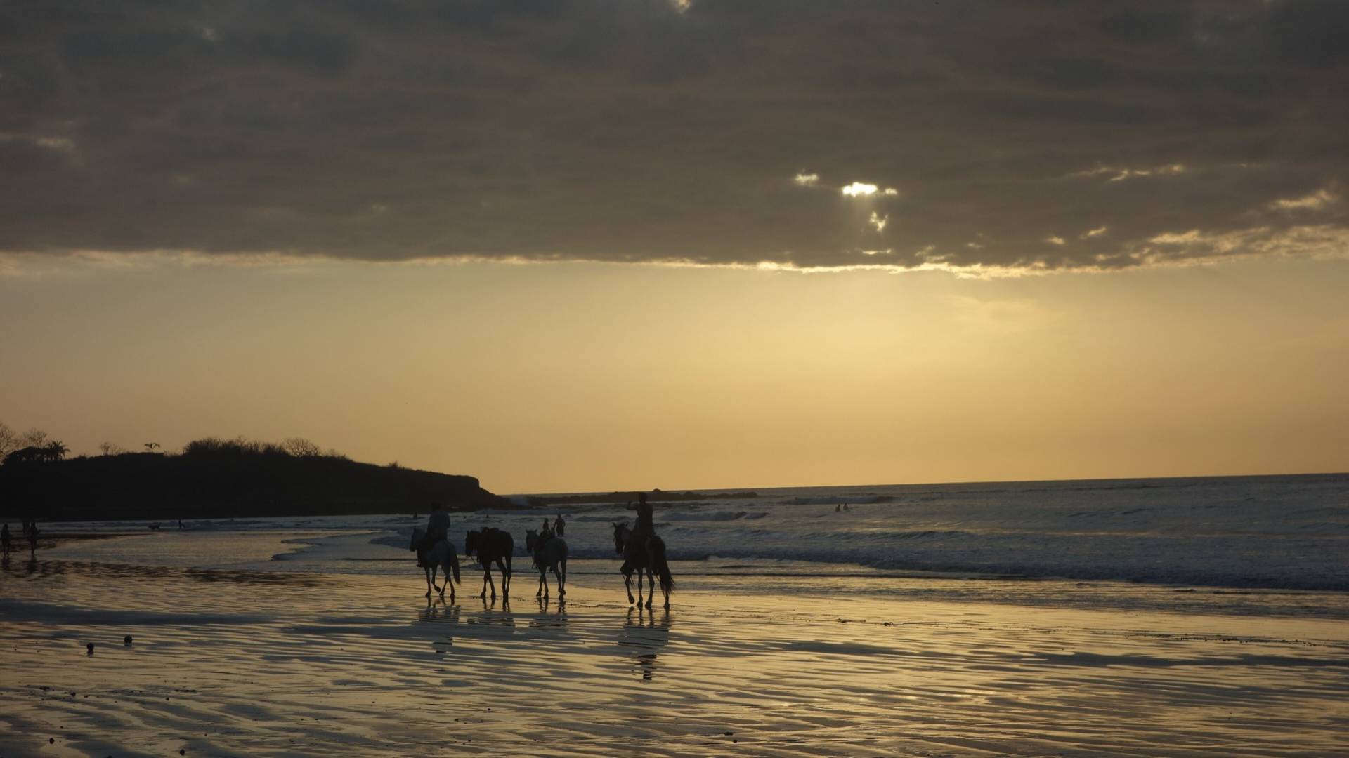 Four riders on horseback silhouetted on the wet sand of Tamarindo beach at sunset, with the sun breaking through dark clouds over the Pacific