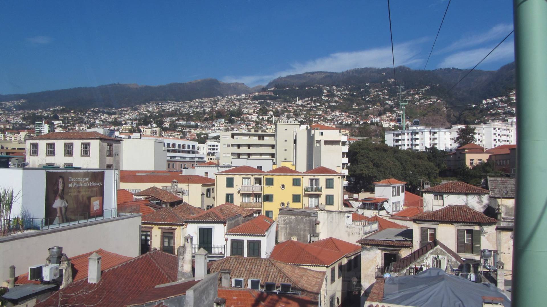 Terracotta rooftops and apartment blocks of Funchal seen from the Monte cable car cabin on a January day, with the cable running up the hill