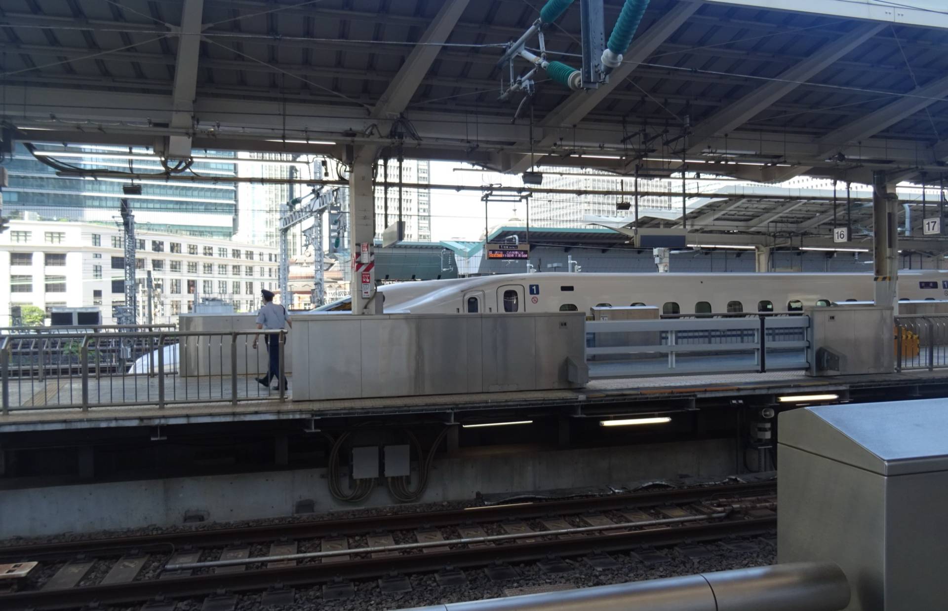 A white N700S Series Shinkansen standing at a Tokyo Station platform behind the platform safety barriers, seen across the tracks from the neighbouring platform, with office towers rising behind the station roof