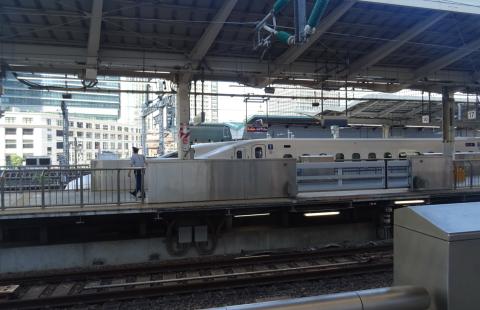 A white N700S Series Shinkansen standing at a Tokyo Station platform behind the platform safety barriers, seen across the tracks from the neighbouring platform, with office towers rising behind the station roof