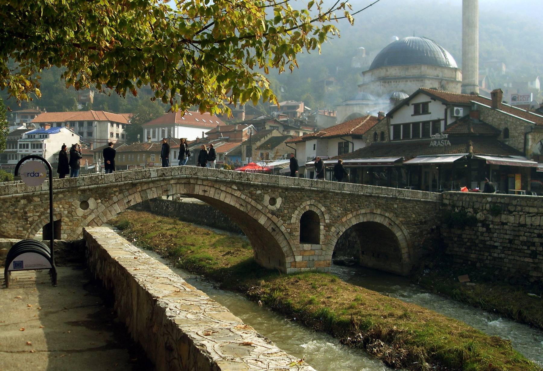 People crossing the arched Stone Bridge over the river in Prizren, with a domed mosque and red-roofed buildings behind