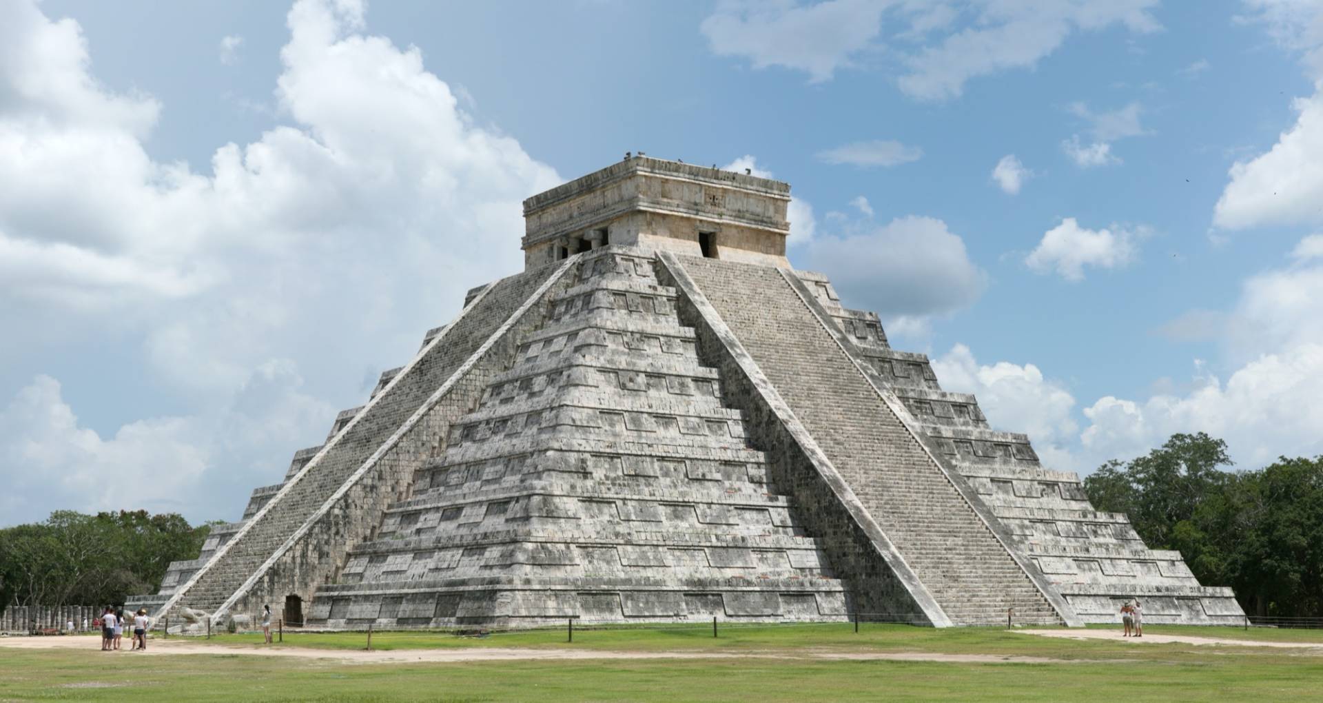 El Castillo pyramid at Chichén Itzá: a nine-tiered stone pyramid with a central staircase and a small temple on top, seen across a mown lawn under a blue sky with white clouds, with a few visitors at its base