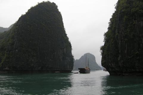 Ha Long Bay on an overcast day: two dark limestone karst towers frame a channel of jade-green water with a wooden junk-style cruise boat and a small rowing boat between them, more karst islands fading into grey haze behind