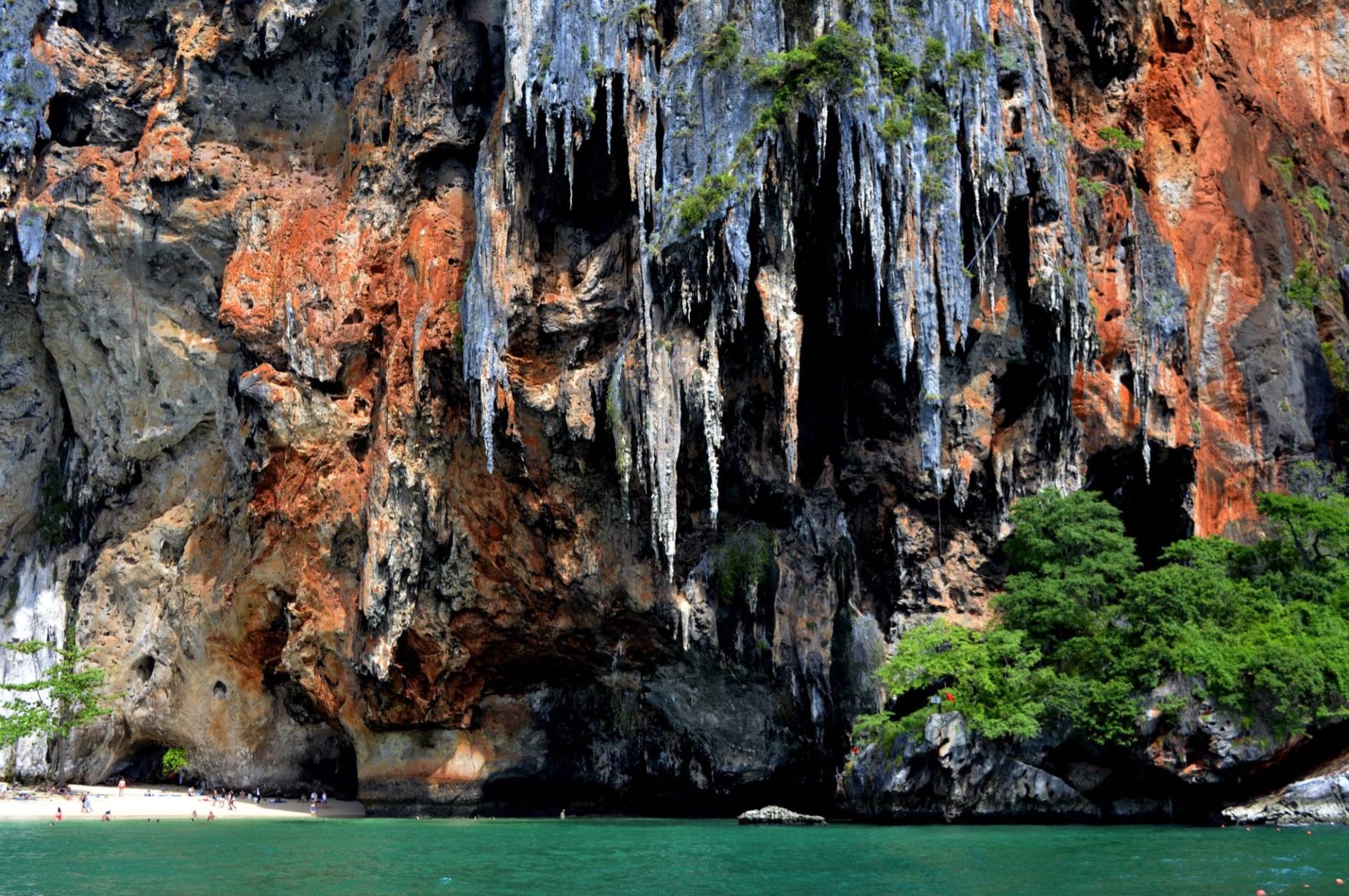 Overhanging limestone cliff with orange-and-grey rock and hanging stalactites above a small beach and turquoise water at Railay, with a few beachgoers dwarfed by the wall
