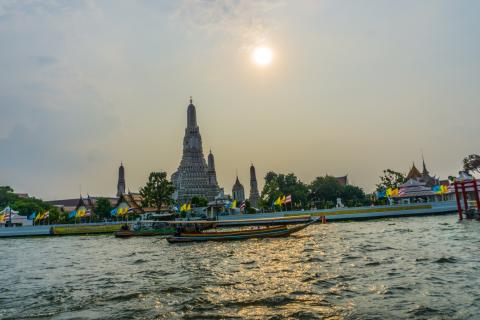 Wat Arun's central prang and smaller spires seen across the Chao Phraya River at sunset, with two long-tail boats passing in the foreground and Thai and royal flags along the riverbank