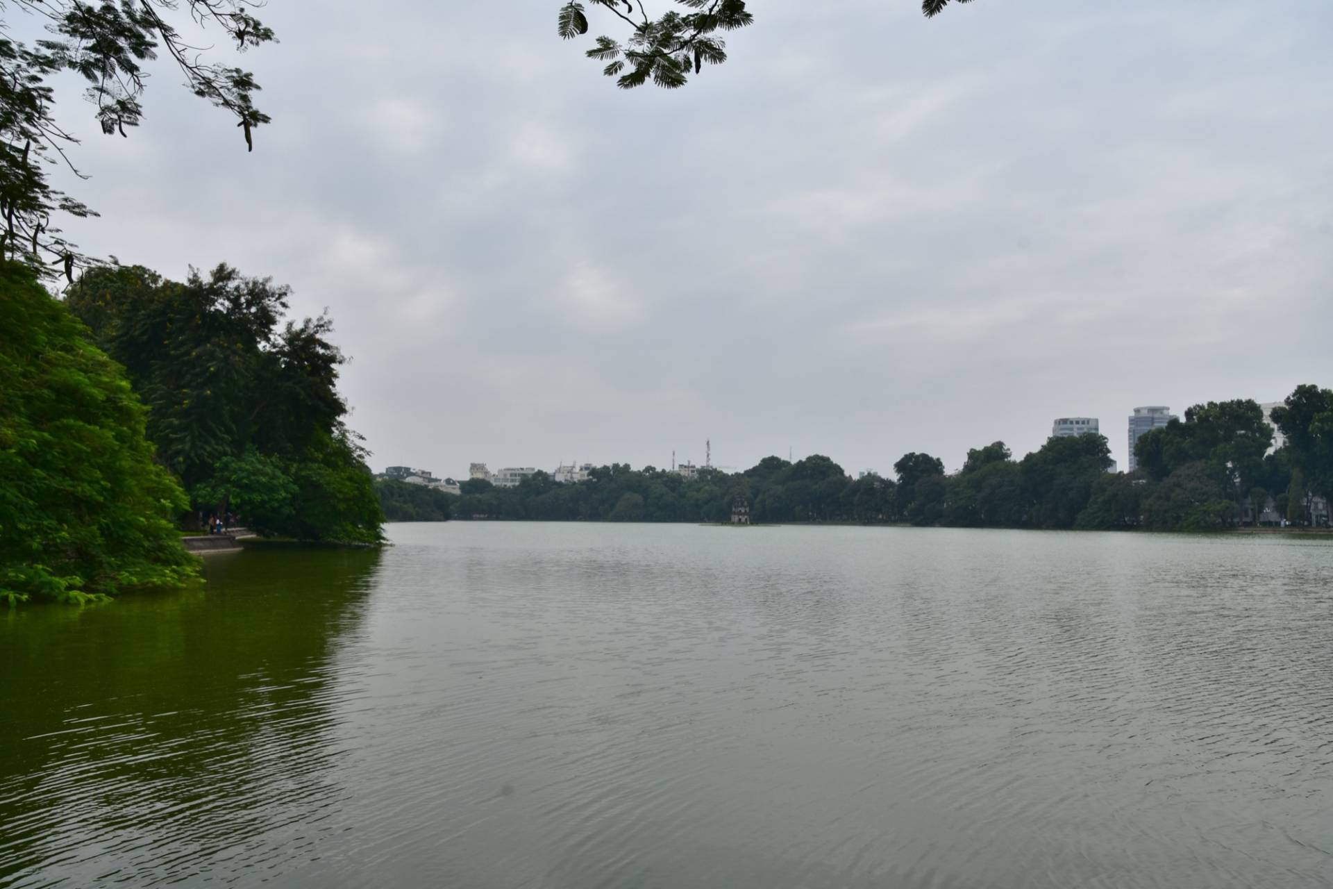 Hoan Kiem Lake in Hanoi under a grey overcast sky: calm green water, tree-lined banks, the small Turtle Tower on its islet in the distance and a few tall buildings on the far shore