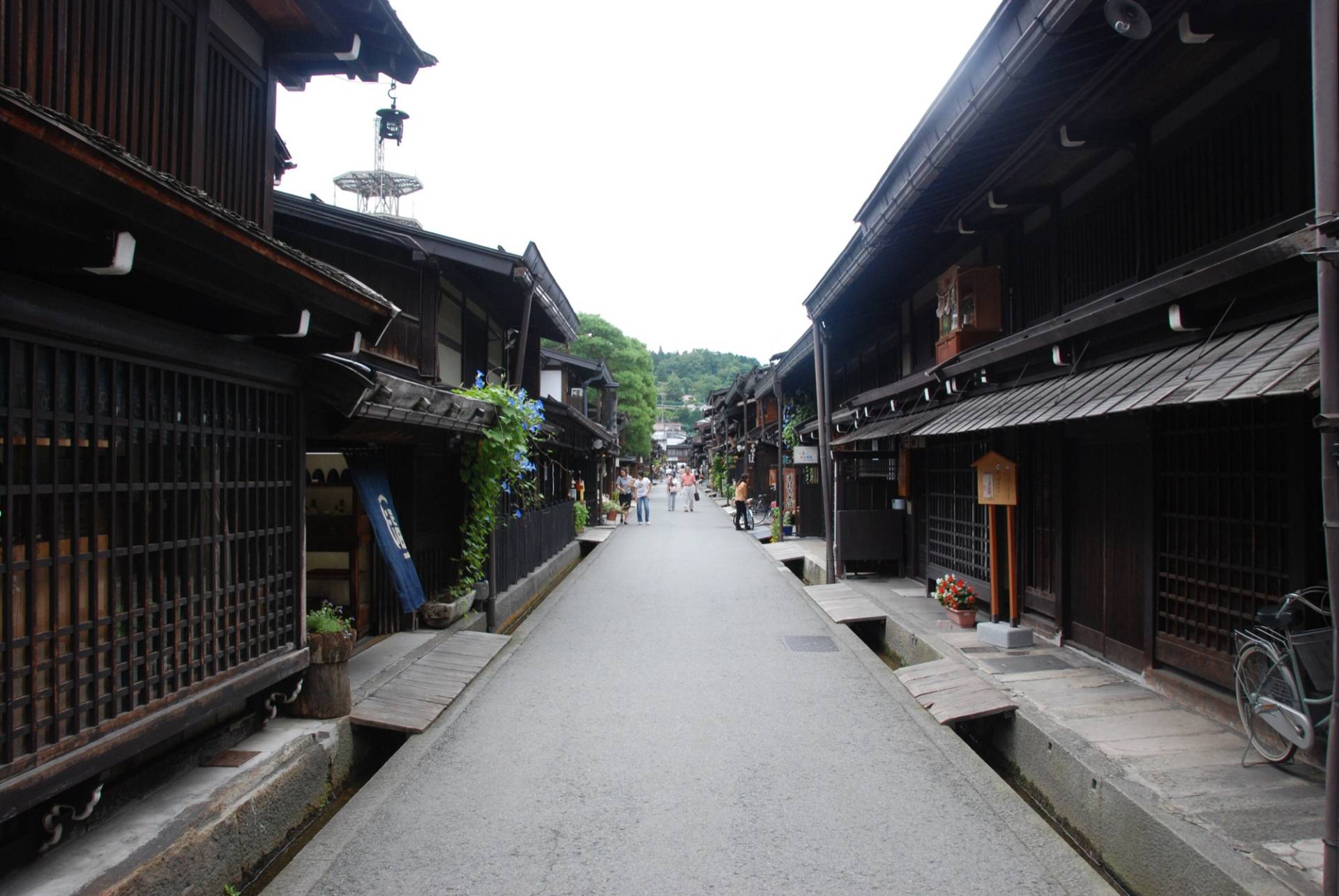 A narrow street in Takayama's Sanmachi district lined on both sides with dark wooden merchant houses, lattice windows, a shop curtain and a bicycle, with a few people walking in the distance