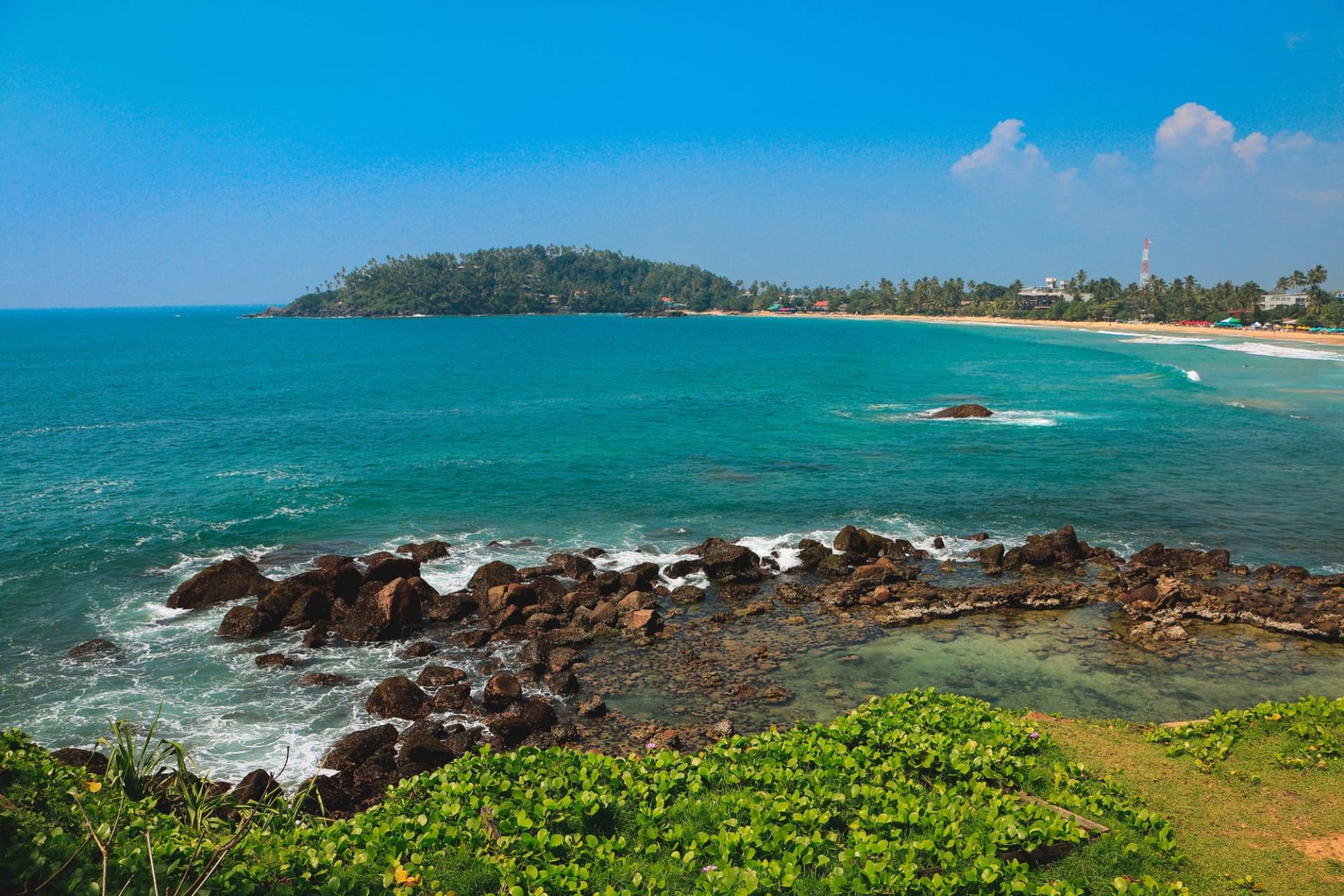 Mirissa bay from a headland: turquoise sea breaking over dark rocks, with the palm-lined beach and the wooded island in the background