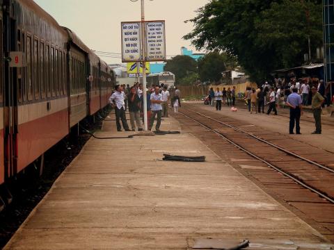 Da Nang railway station platform: a long Vietnam Railways passenger train in red, white and green livery on the left, groups of passengers waiting on the concrete platform beside bilingual safety signs, tracks curving away to the right