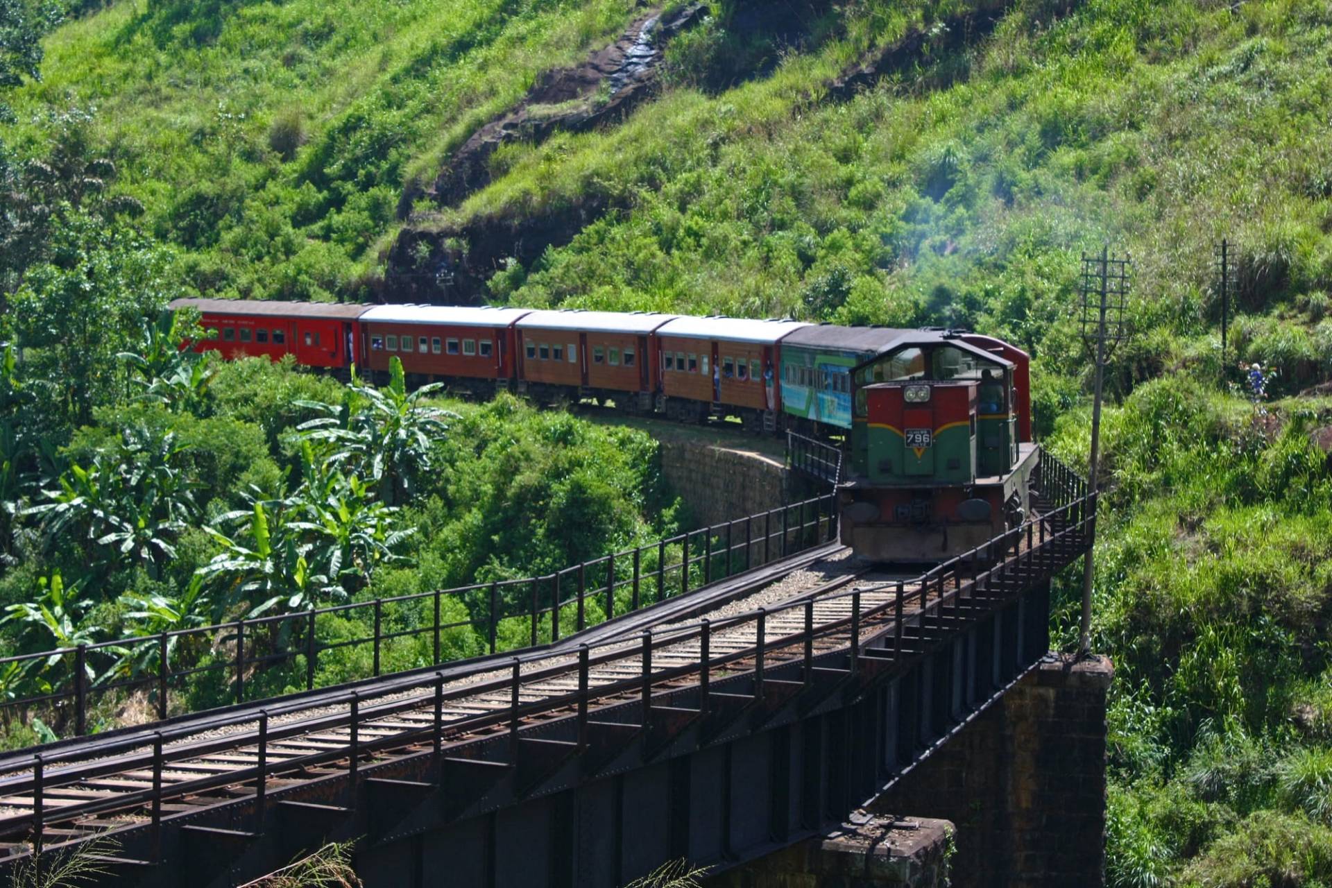 A red and green Sri Lanka Railways train crossing an iron bridge on a curve as it approaches Demodara station, with green hillside behind