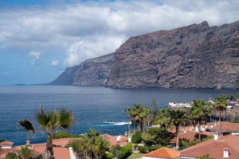 Dark sea cliffs of Los Gigantes rising from the blue sea with palms and tiled roofs in the foreground