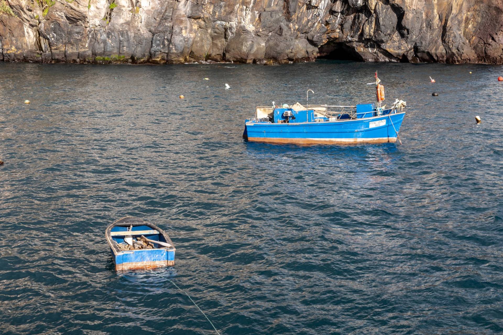 Two small blue fishing boats moored on dark water below the cliff at Câmara de Lobos harbour