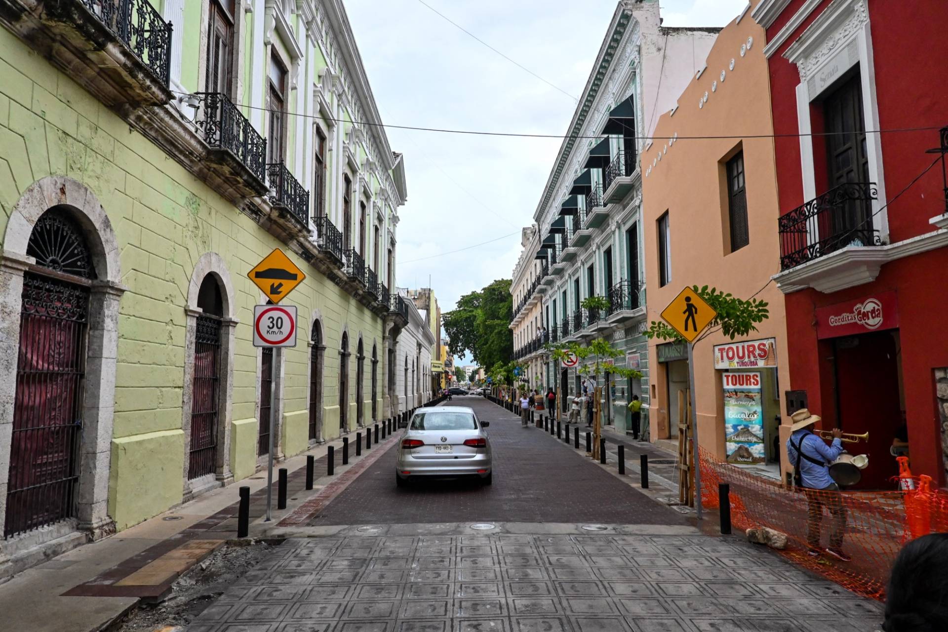 Calle 60 in the centre of Mérida, Yucatán: a narrow street of two-storey colonial buildings painted lime green, teal, orange and red with wrought-iron balconies, a single car and a musician on the right-hand pavement