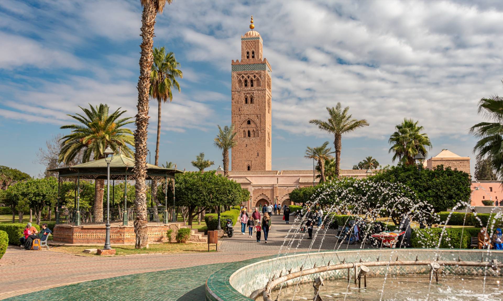 The Koutoubia minaret in Marrakech rising above palm trees and a green-tiled fountain in the foreground, people walking in the park under a sky of broken cloud